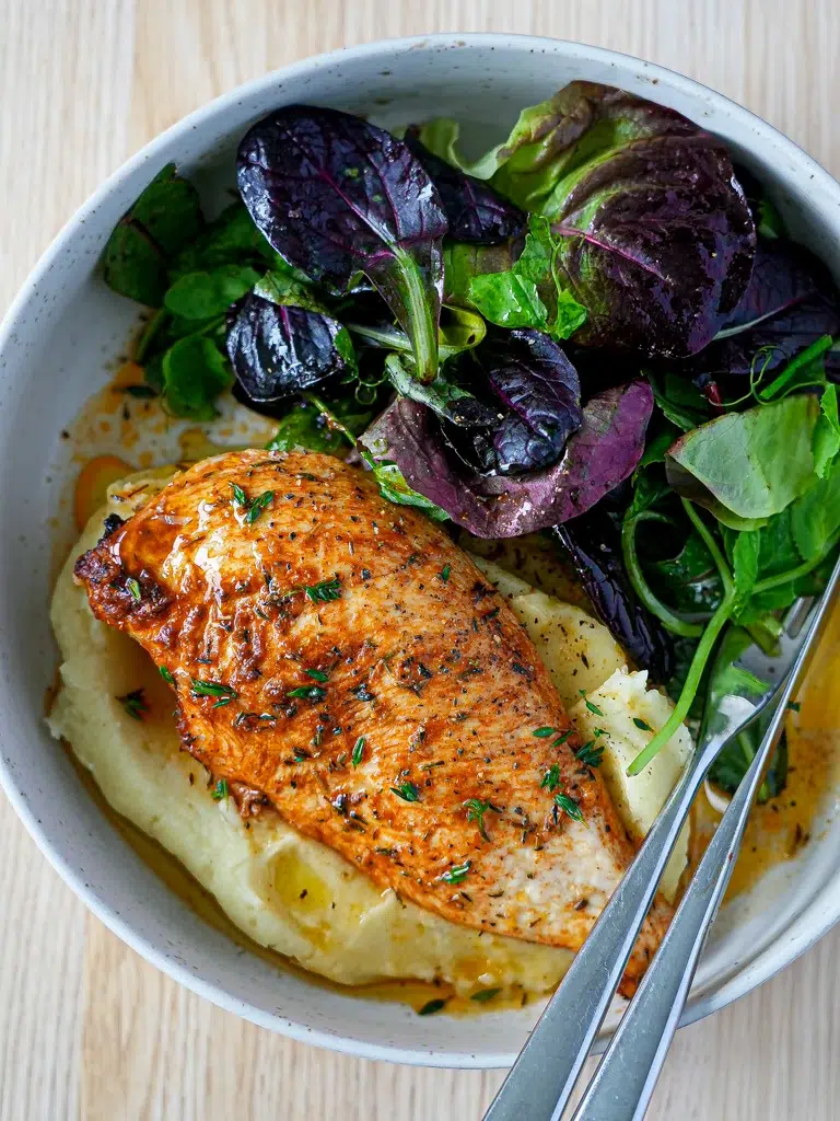 A bowl of Baked garlic chicken breast cooked at high heat for tenderness and juiciness, served with mashed potato and fresh herbs