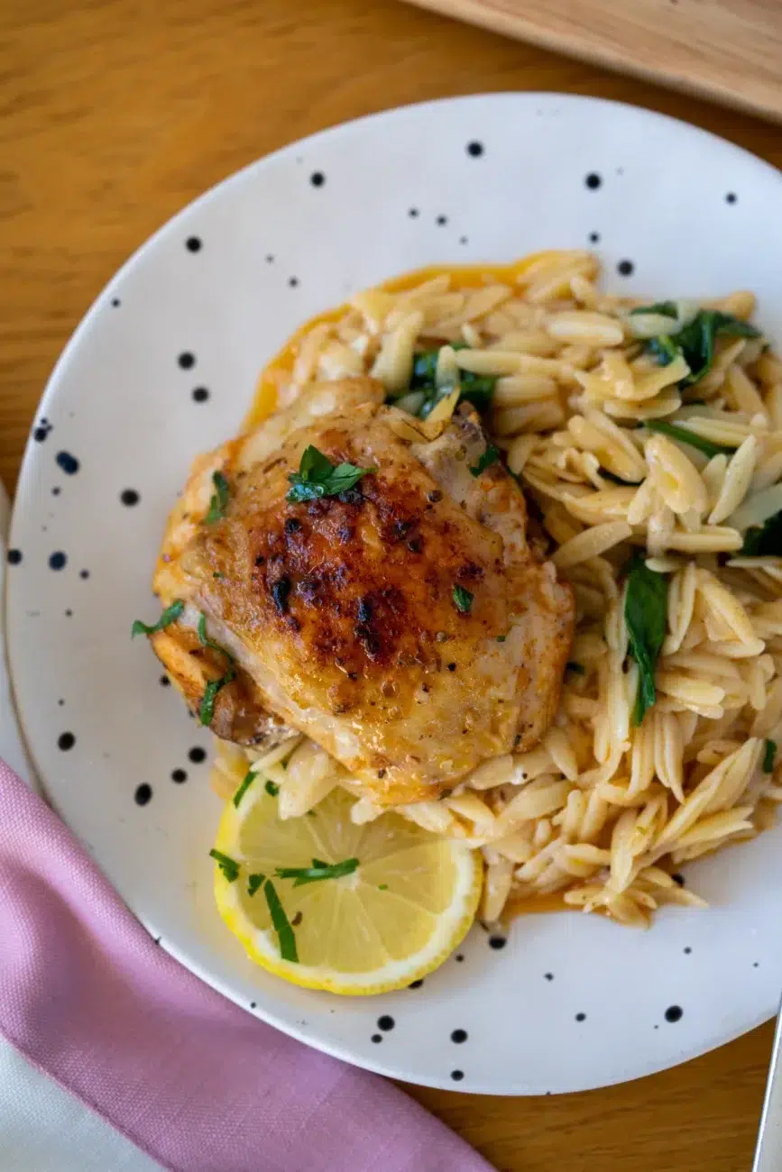 Crispy one pan Greek chicken served on a bed of creamy risoni pasta with spinach and garnished with fresh parsley and lemon slice on a speckled plate.