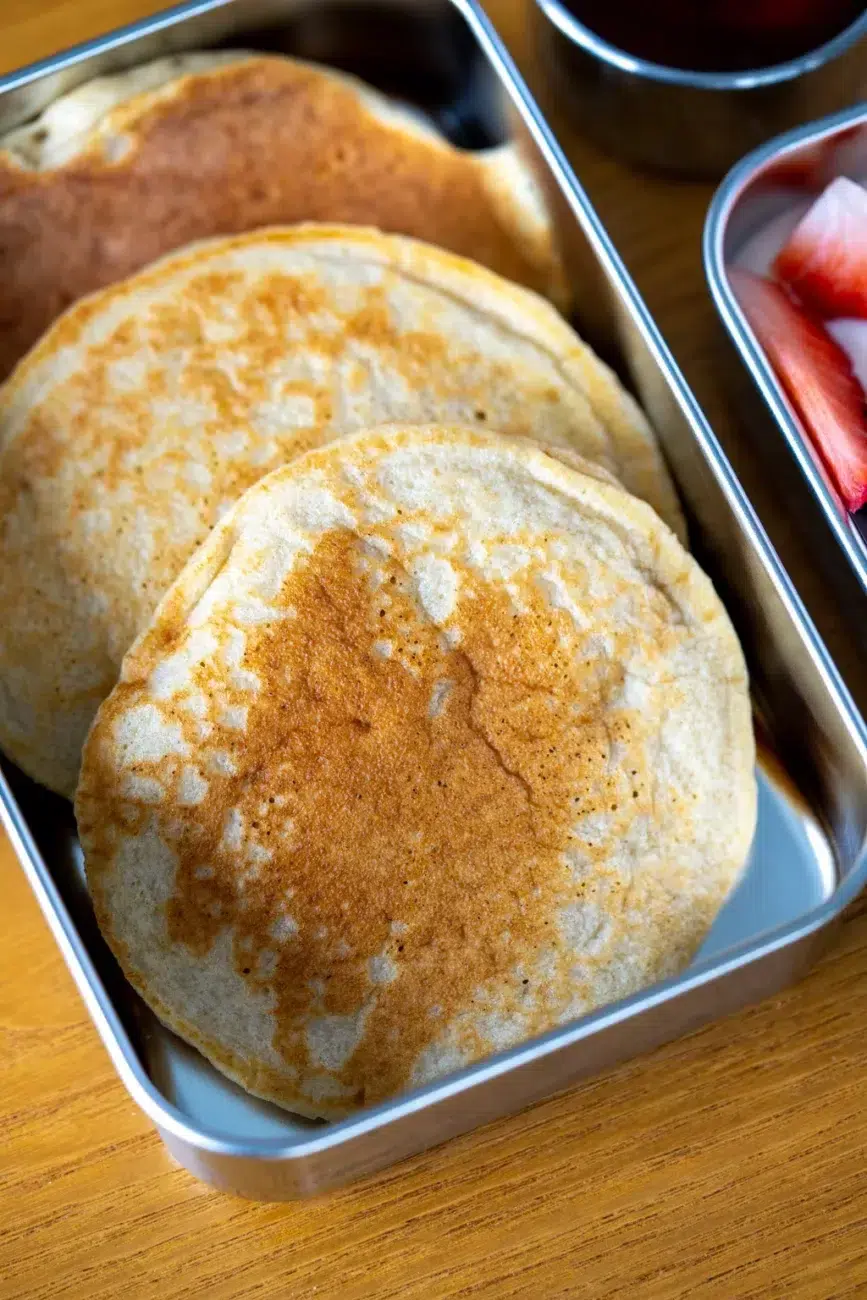 Close-up side view pancakes in stainless steel lunchbox with fruit and maple syrup on side