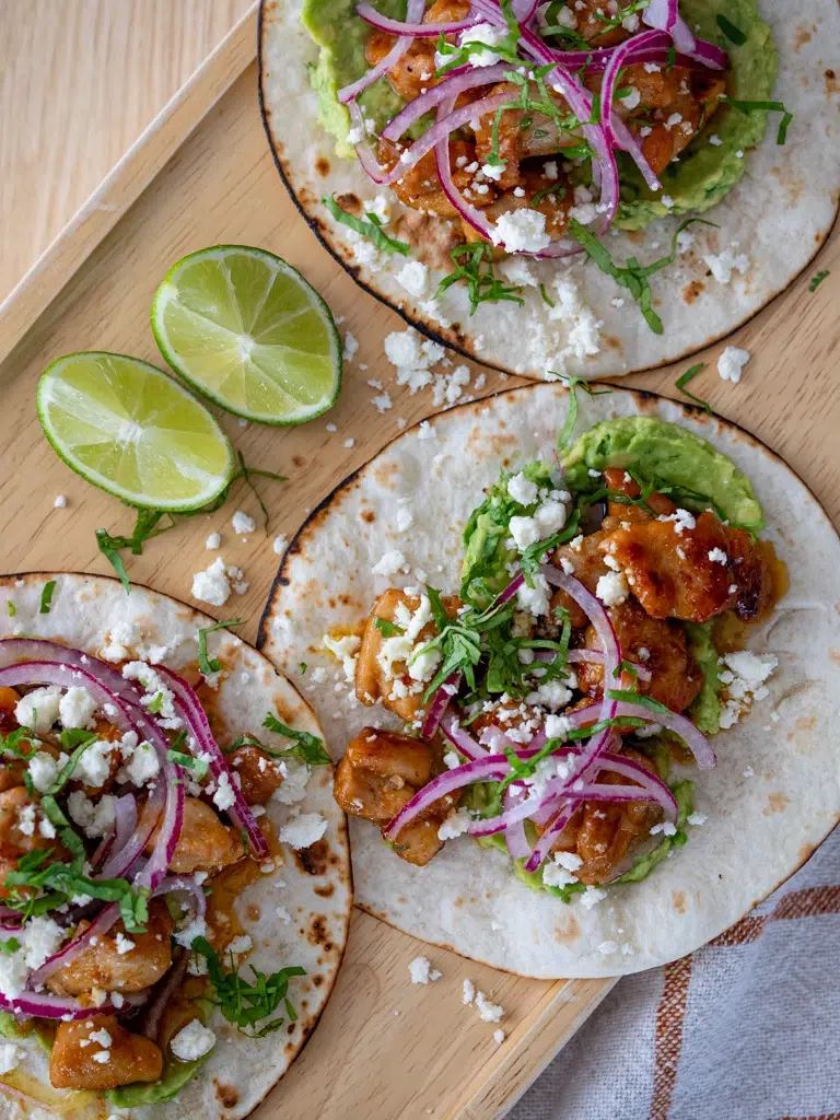Three sticky lime honey glazed chicken tacos on toasted tortillas with guacamole, pickled red onions, crumbled cheese, and fresh herbs, served with lime wedges on a wooden tray.